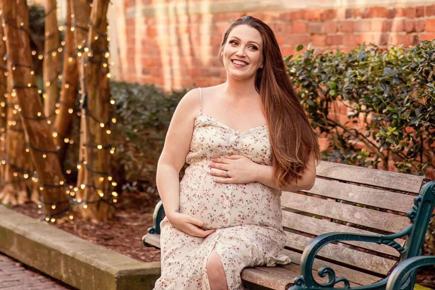 Pregnant woman in floral dress sitting on bench with string lights in background.