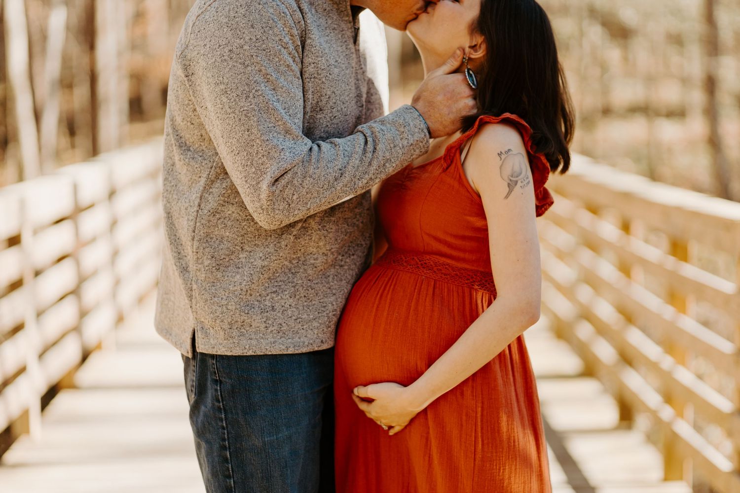 Couple sharing a tender kiss on a wooden bridge, pregnant woman in rust orange maternity dress holding her baby bump.