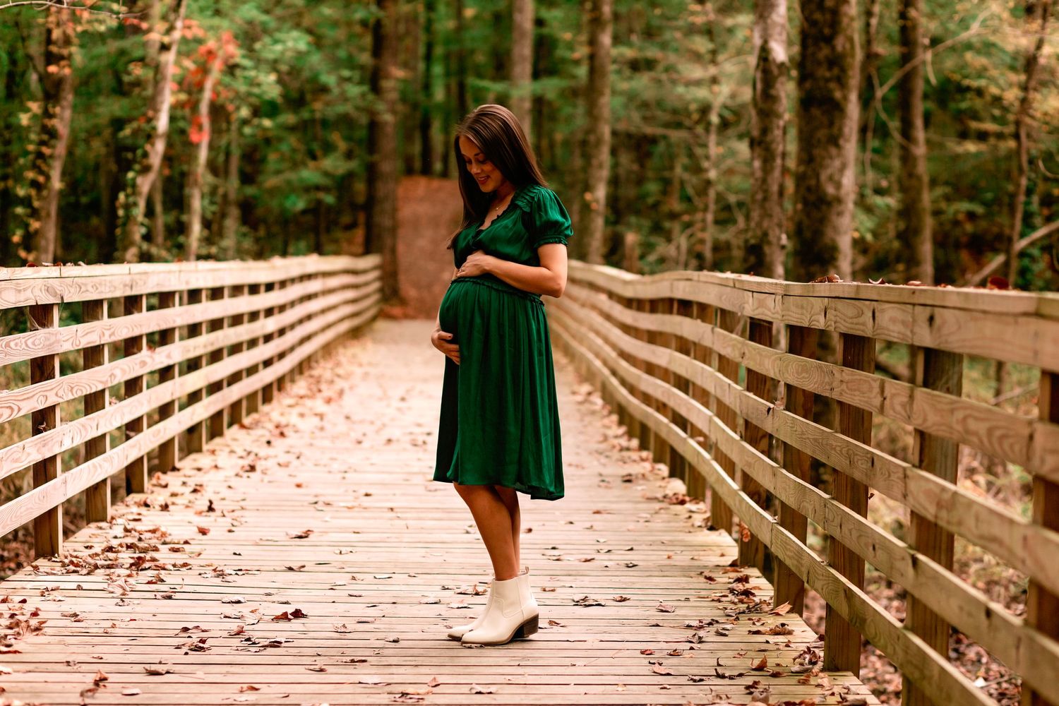 Pregnant woman in emerald green dress cradling baby bump on wooden bridge in forest.