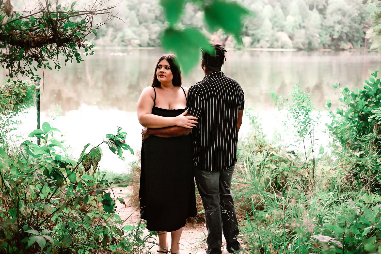 Pregnant woman in black dress standing with partner near a serene lake surrounded by lush green foliage.