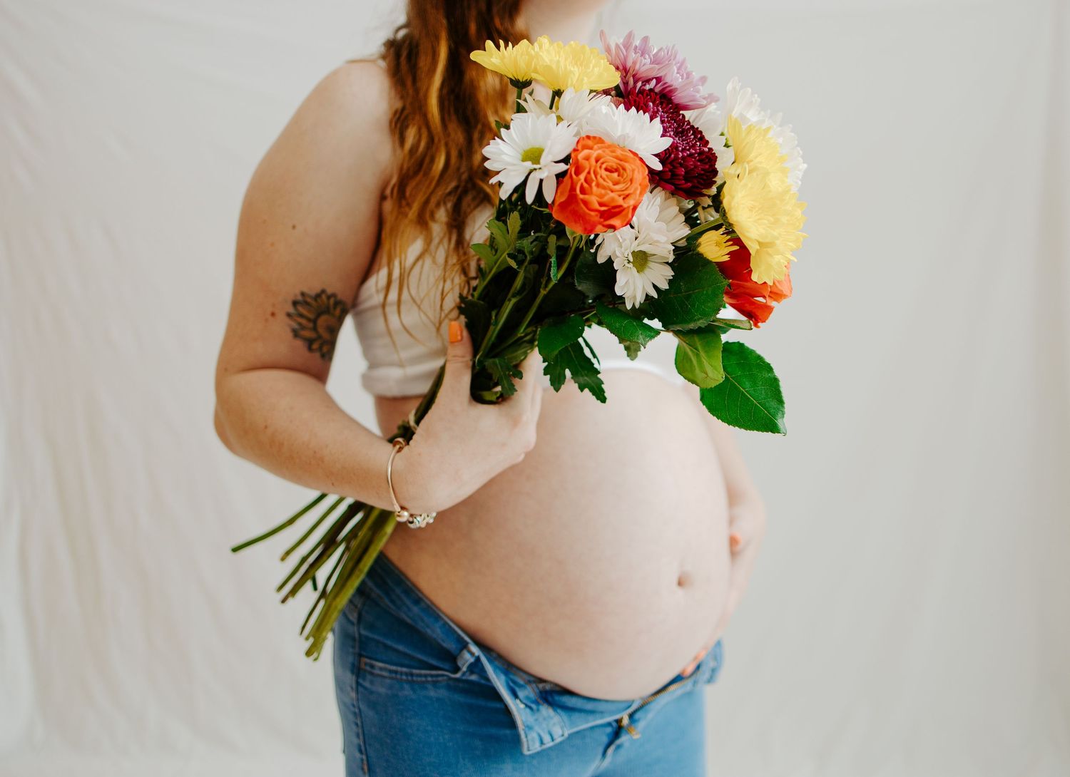 Pregnant woman in crop top and jeans holding vibrant bouquet of colorful flowers against white background.
