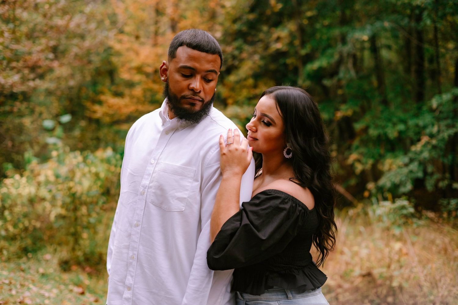 Couple posing together in an autumn forest, woman in black off-shoulder top leaning on man.