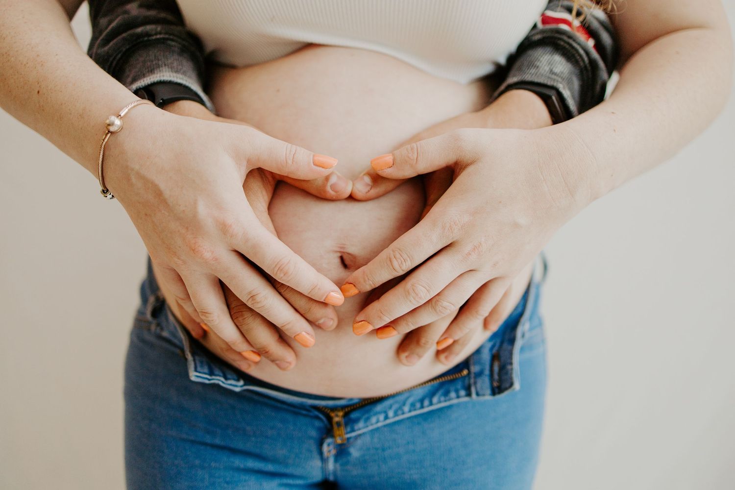 Close-up of couple making heart shape with hands on pregnant belly, woman wearing orange nail polish and gold bracelet.