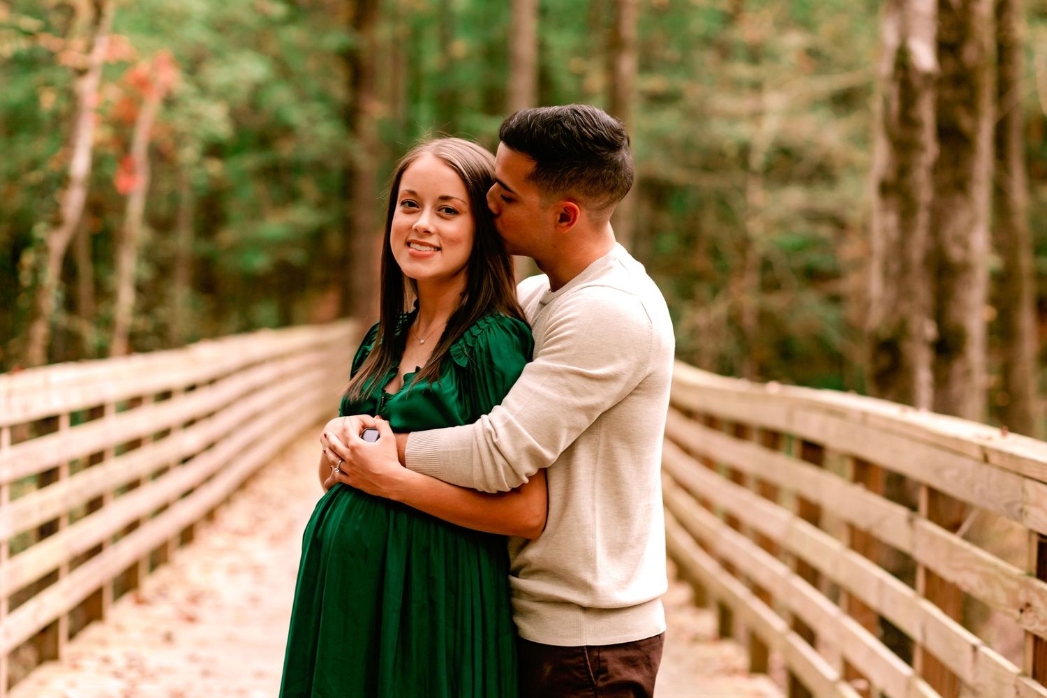 Pregnant woman in green dress embraced by partner on wooden bridge in autumn forest.