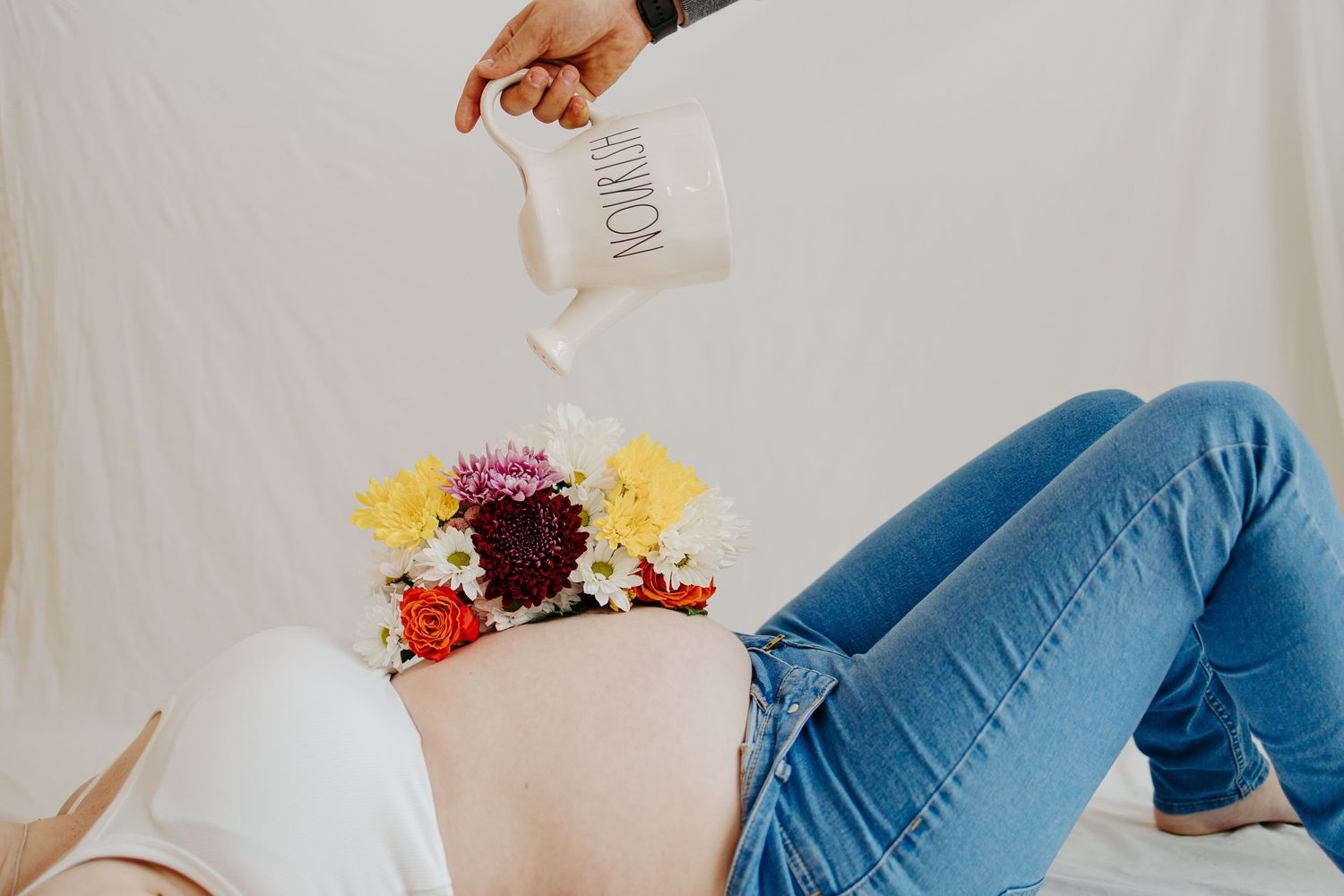 Creative maternity photo of pregnant woman lying down with flowers on belly while someone pours from a Rae Dunn mug.