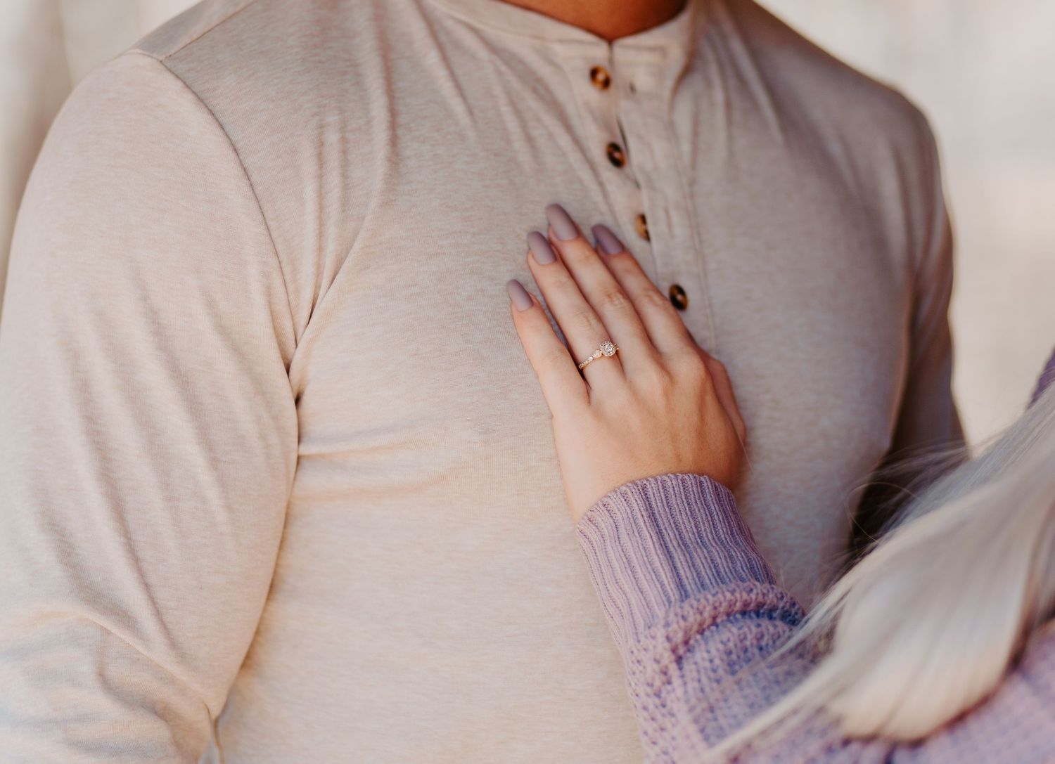 Close-up of womans hand with engagement ring resting on mans cream henley shirt.