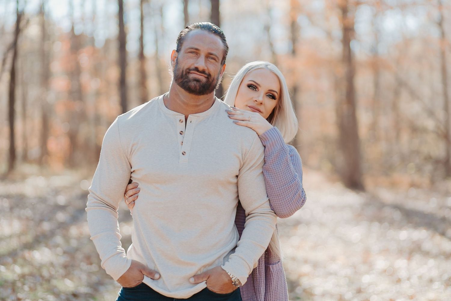 Couple posing together on a forest path in autumn, man in white henley with woman in lavender top leaning on his shoulder.
