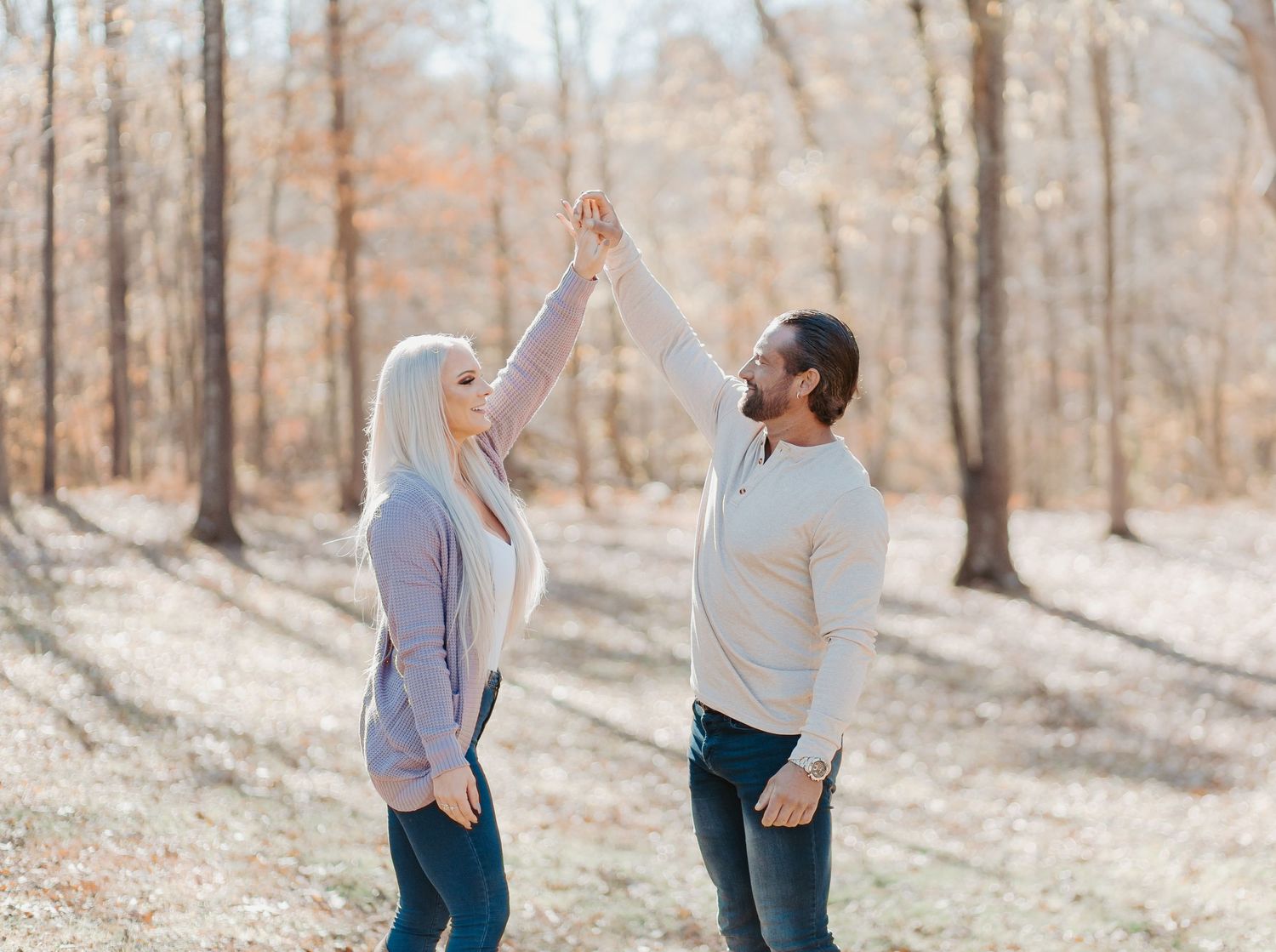 Couple dancing outdoors in autumn forest, raising hands together in a playful spin among fall foliage.