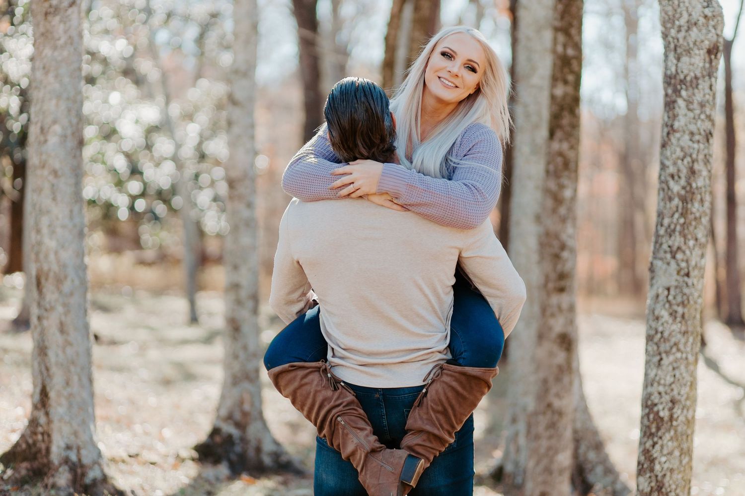 Woman smiling while being carried piggyback by man among bare trees in an outdoor autumn setting.