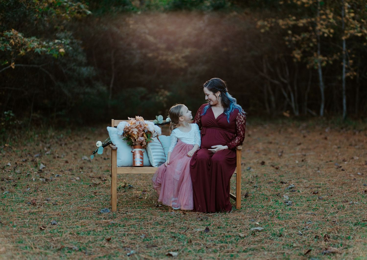Pregnant woman in burgundy lace gown sitting with young girl in pink dress on garden bench.