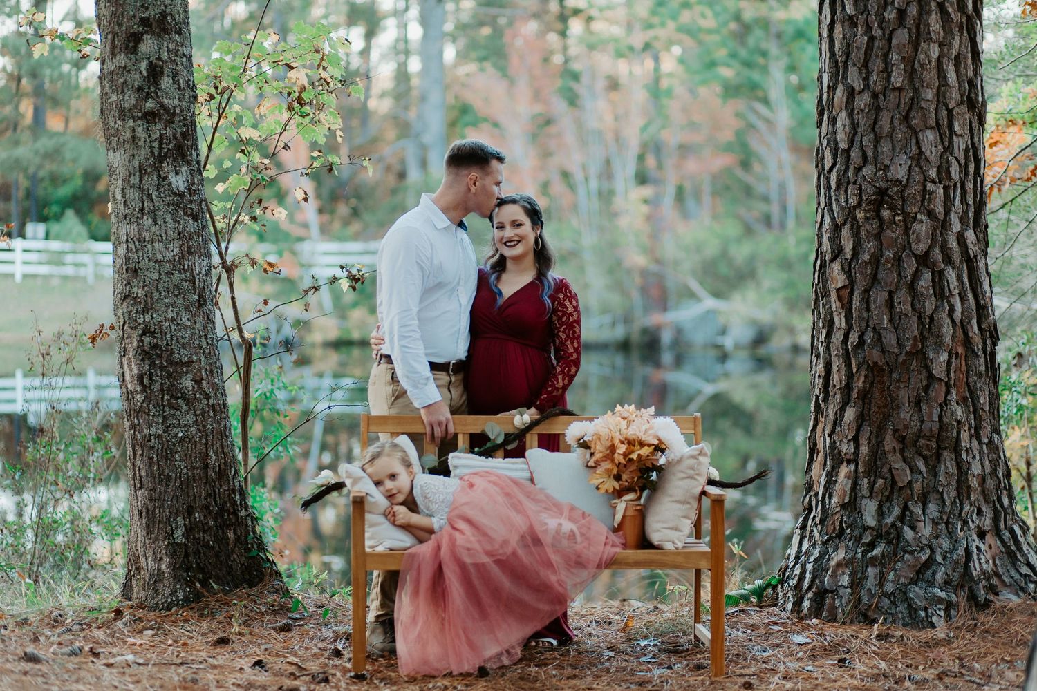Pregnant couple sharing a kiss beside a wooden bench with floral decor among tall trees.