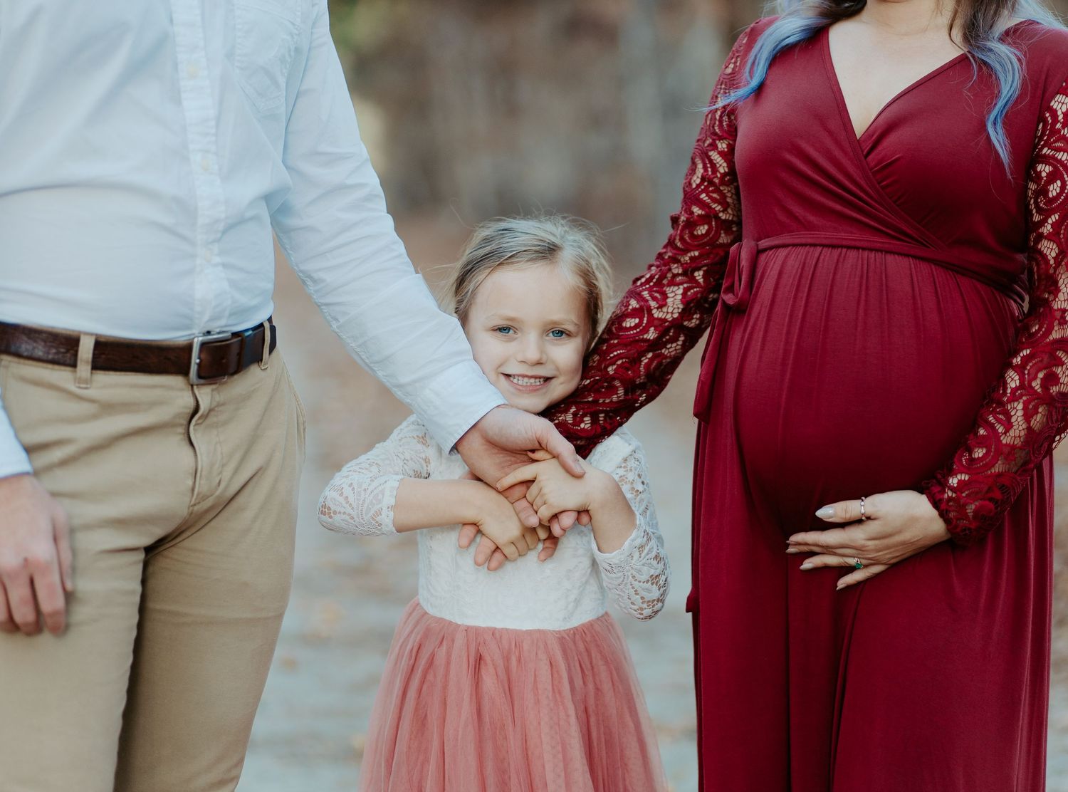 Pregnant woman in red lace dress holding hands with young daughter and partner outdoors.