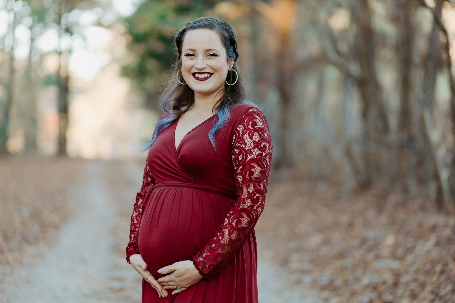 Smiling pregnant woman in burgundy lace maternity gown posing on a tree-lined autumn path.