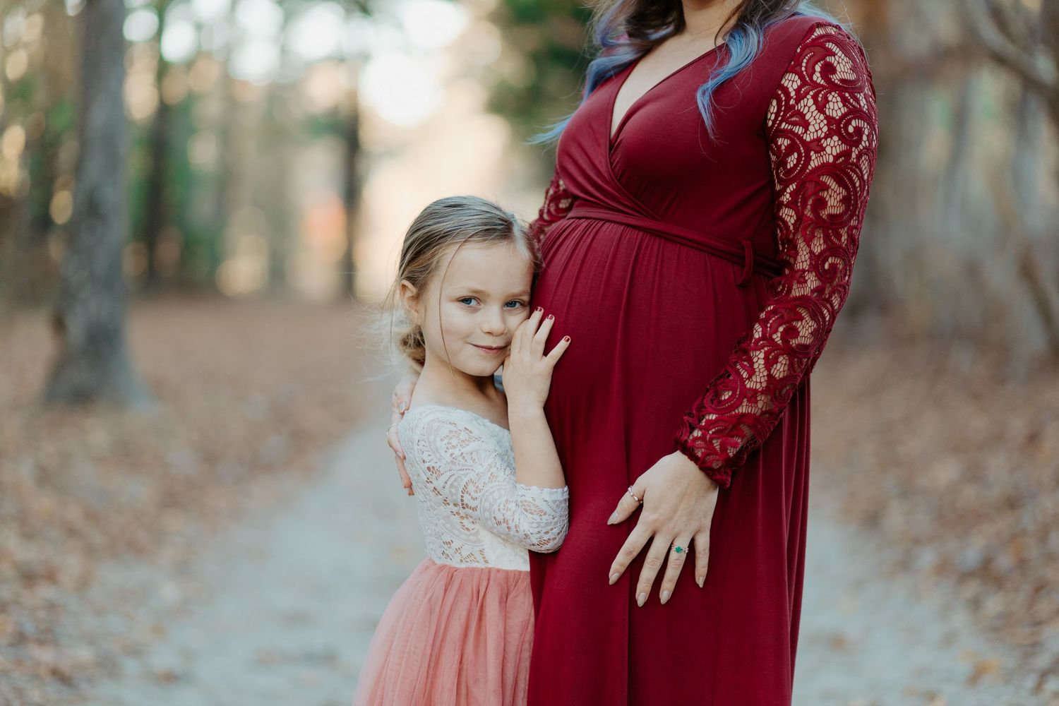 Young girl in pink tulle dress leaning against pregnant mothers burgundy gown outdoors.