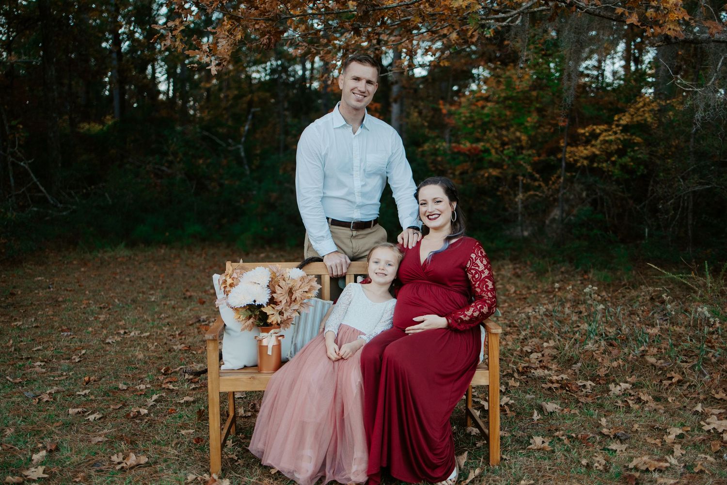 Family maternity portrait with pregnant woman in red dress seated on bench in autumn woods.