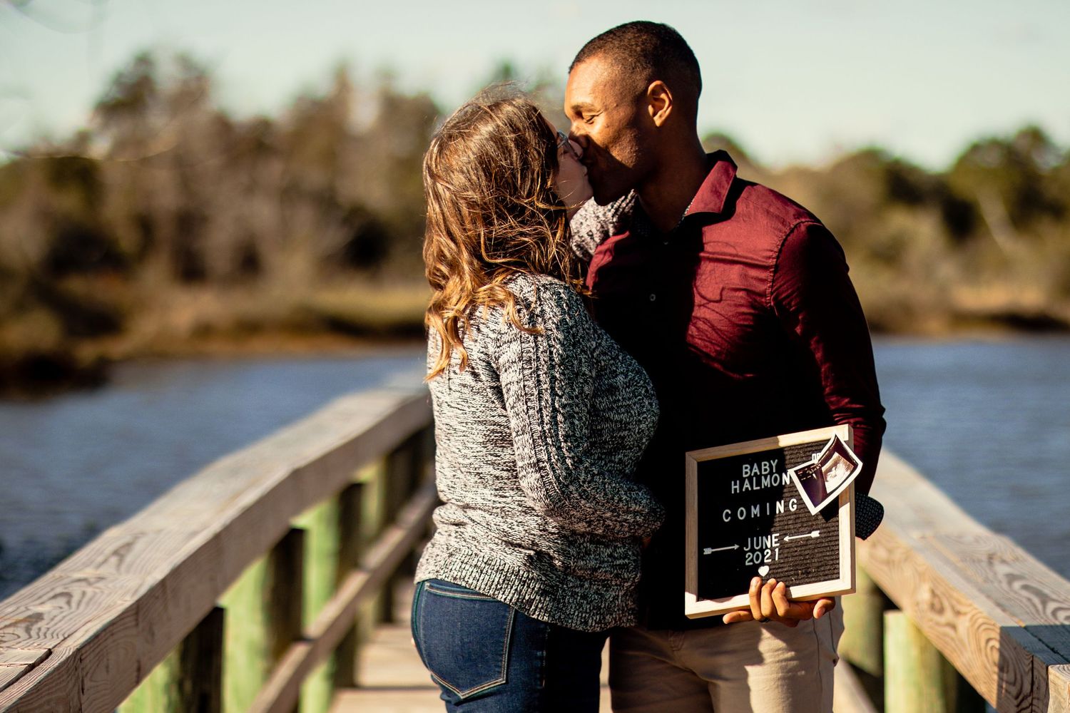 Couple kissing on bridge holding baby announcement sign with ultrasound photos.
