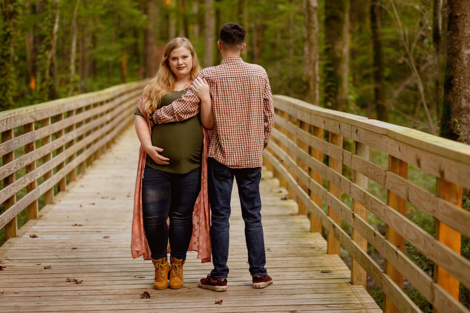 Pregnant couple standing on wooden bridge in forest, woman cradling baby bump during maternity photoshoot.