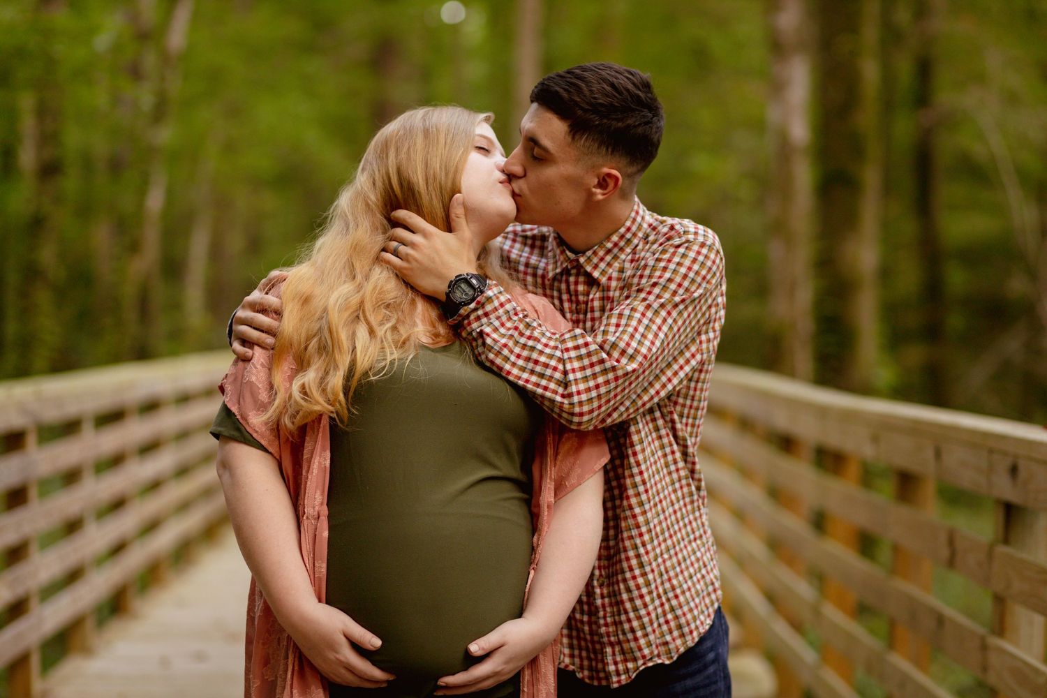 Expecting couple sharing a kiss on a wooden bridge surrounded by lush green trees during maternity session.