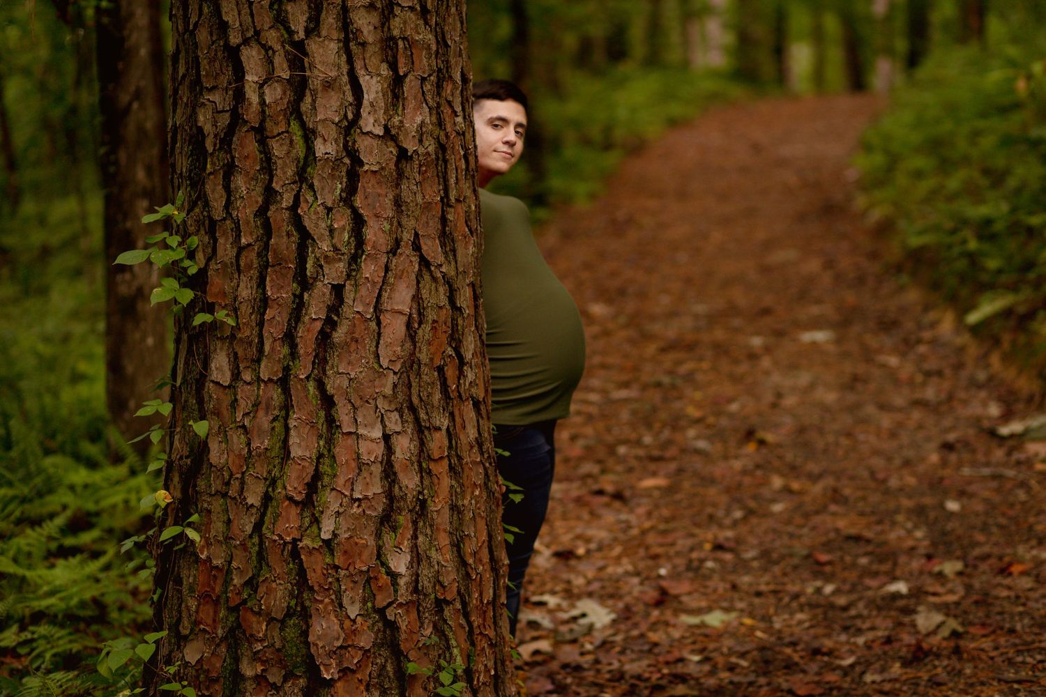Pregnant woman in green top peeks around a large tree trunk on a forest path covered in autumn leaves.