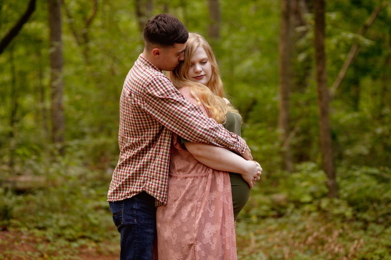 Couple embracing tenderly on a wooded forest path, man in plaid shirt hugging pregnant woman in pink dress.