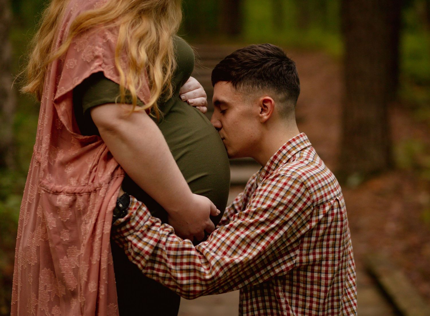 Man kneeling to kiss pregnant belly of woman in pink dress on a forest path surrounded by green trees.