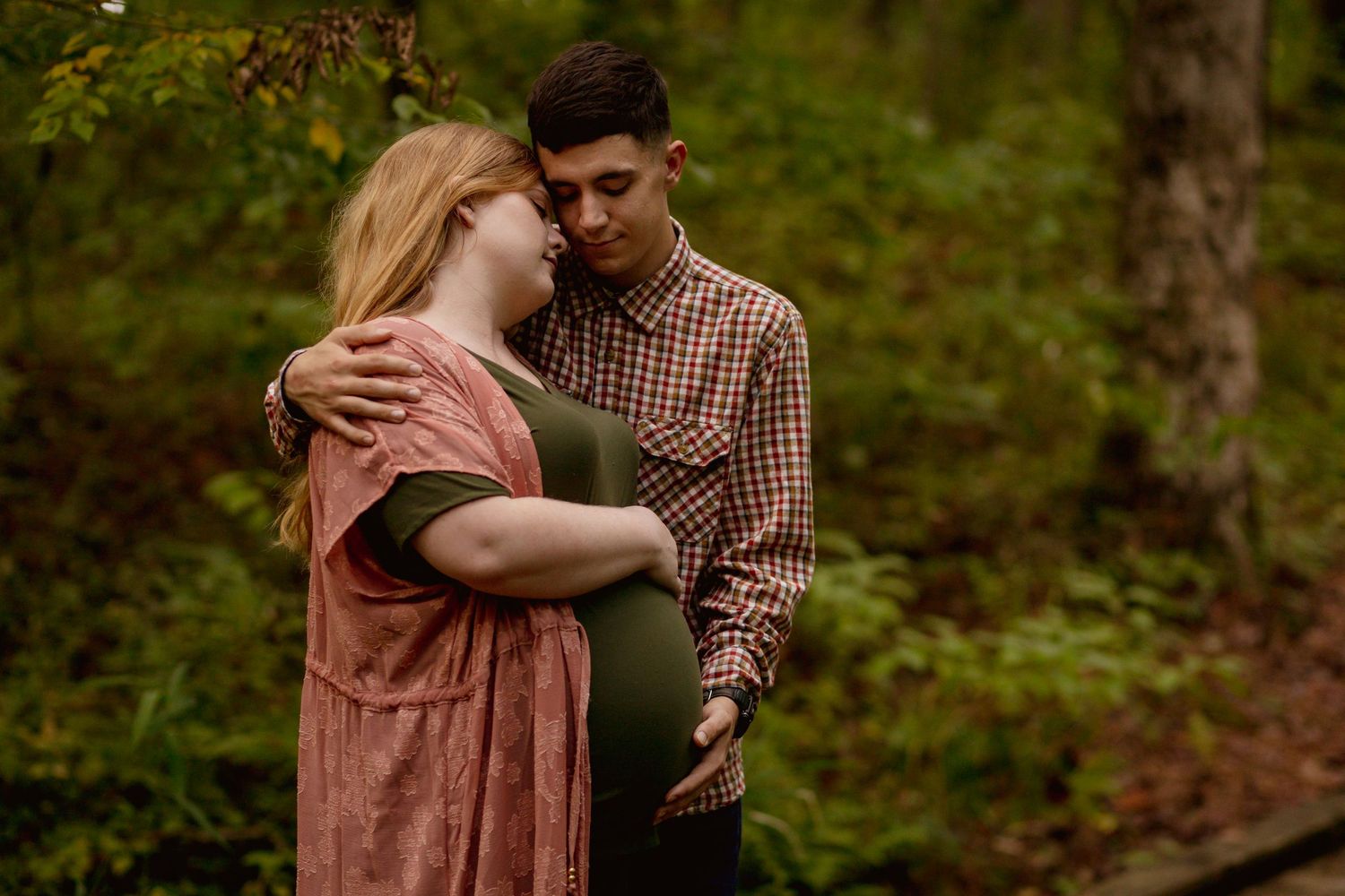 Expecting couple sharing an intimate moment on a forest path, man in plaid cradling pregnant womans belly.
