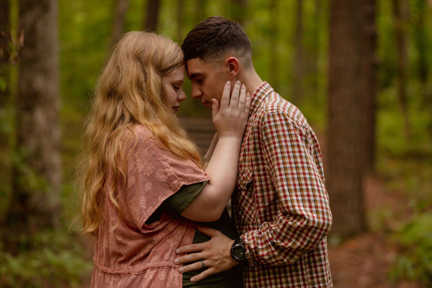 Couple sharing a tender forehead-to-forehead moment on a green forest path, man gently holding womans face.