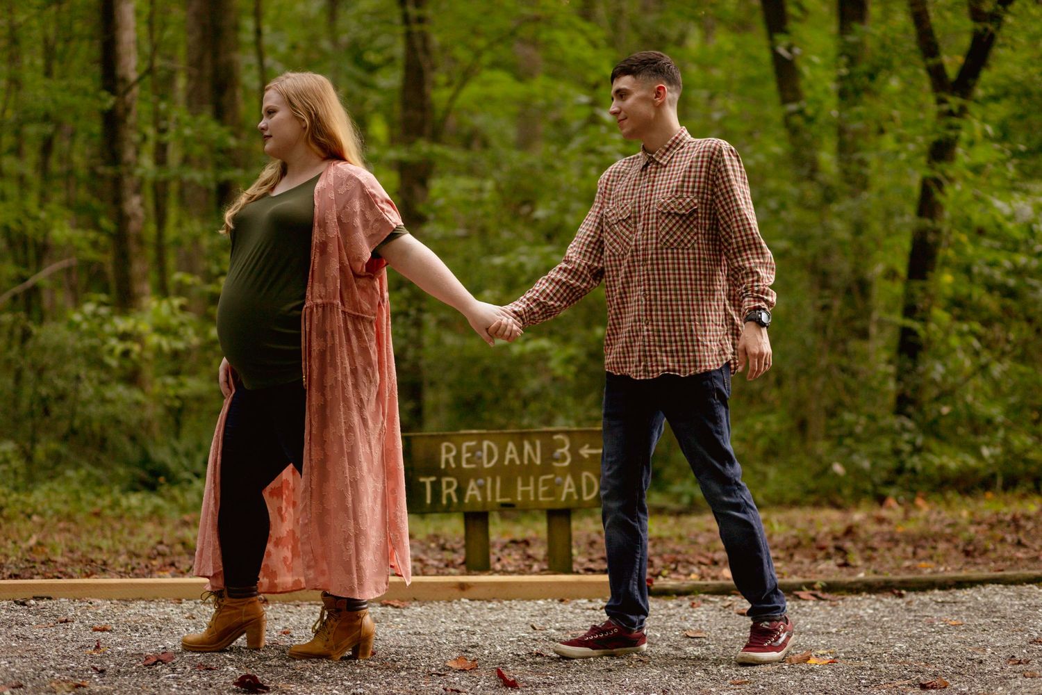 Pregnant couple holding hands walking past a Redan B Trailhead sign surrounded by lush green forest.