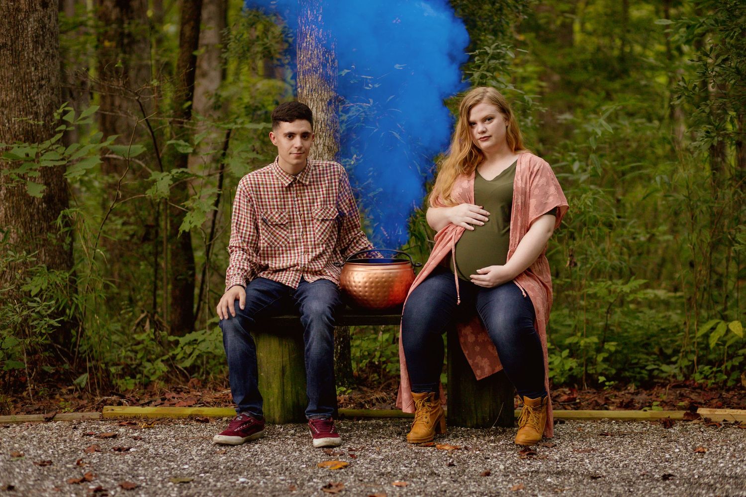 Couple sitting on a bench with blue gender reveal smoke in a lush green forest setting.