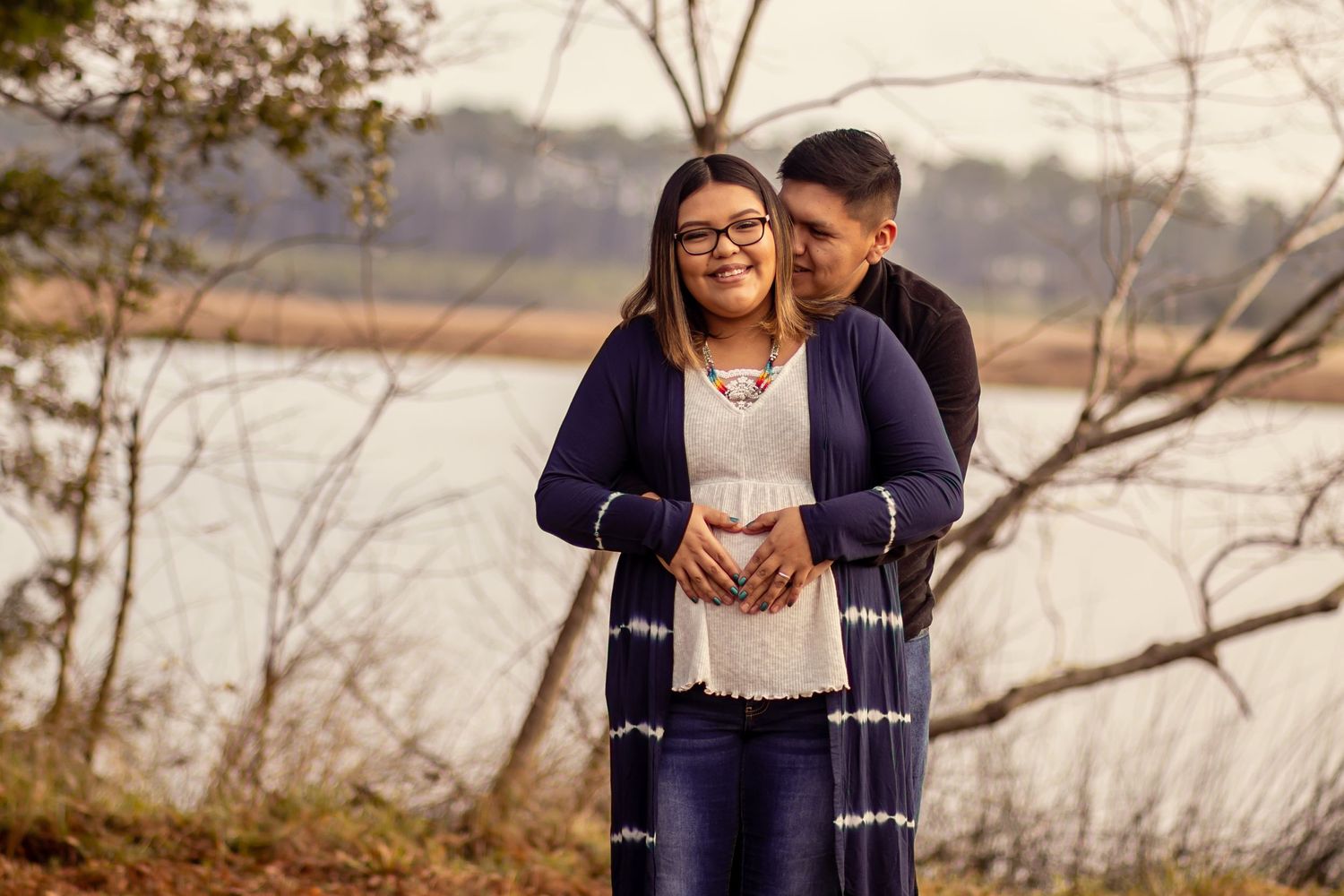 Happy pregnant couple embracing near lakeside, woman in navy tie-dye cardigan smiling during maternity session.