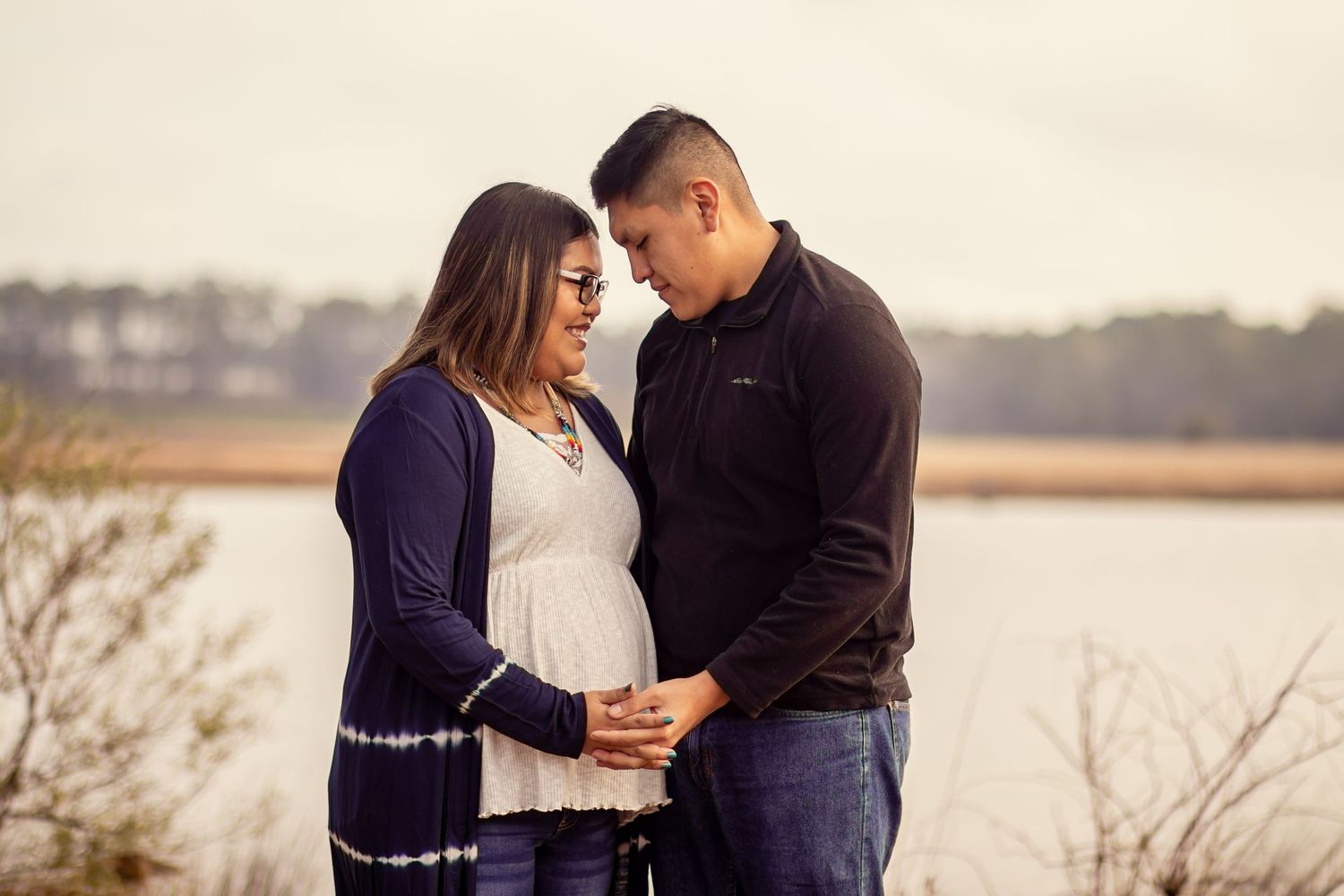 Couple sharing tender maternity moment by lakeside, woman in navy cardigan cradling baby bump outdoors.