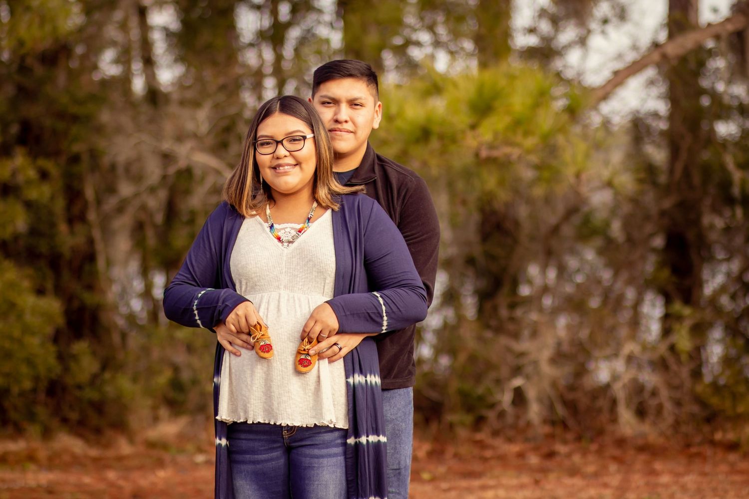 Smiling couple posing outdoors in woods, woman wearing glasses and navy cardigan held from behind by partner.