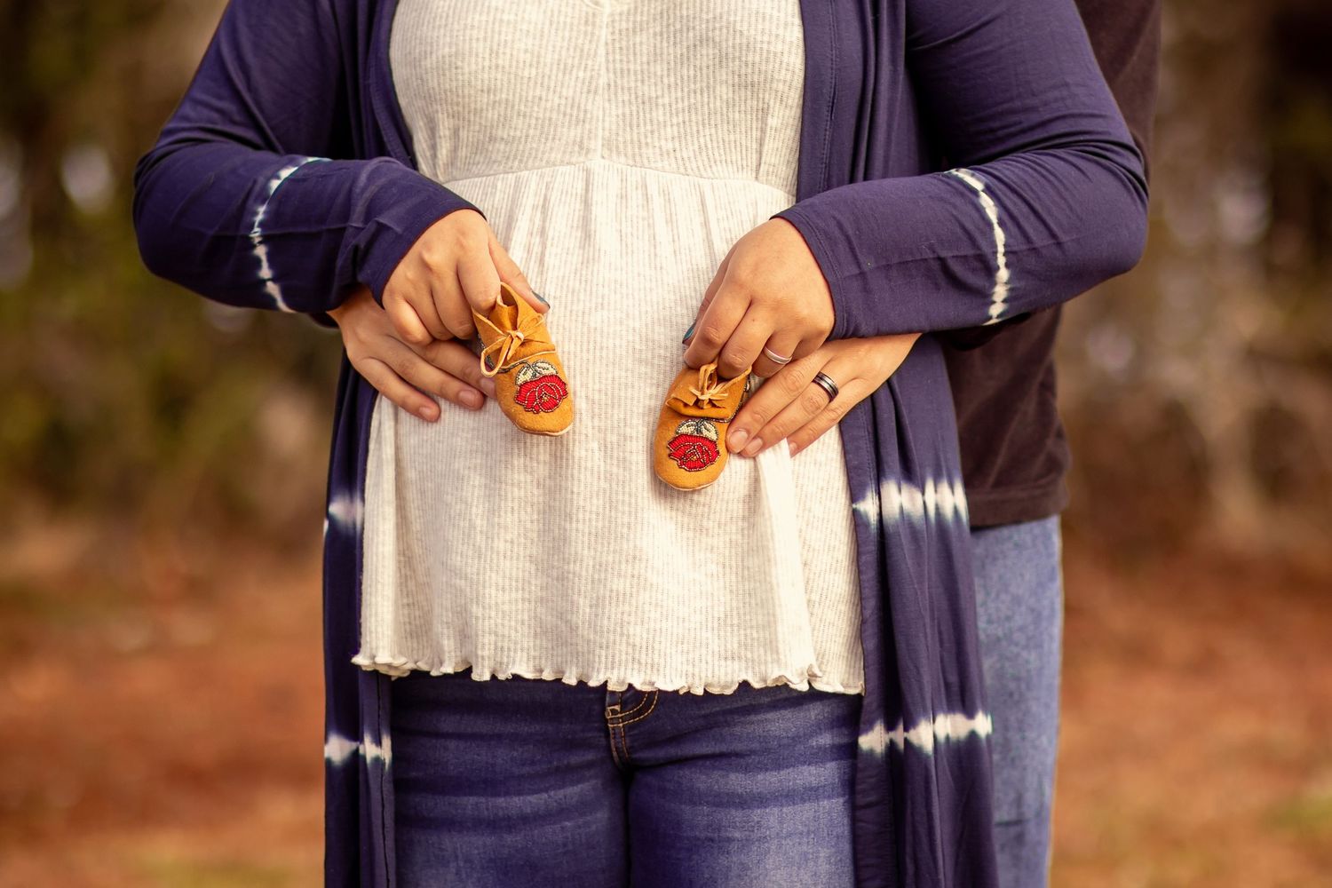 Close-up of pregnant belly with couple holding tiny orange baby shoes during outdoor maternity photoshoot.
