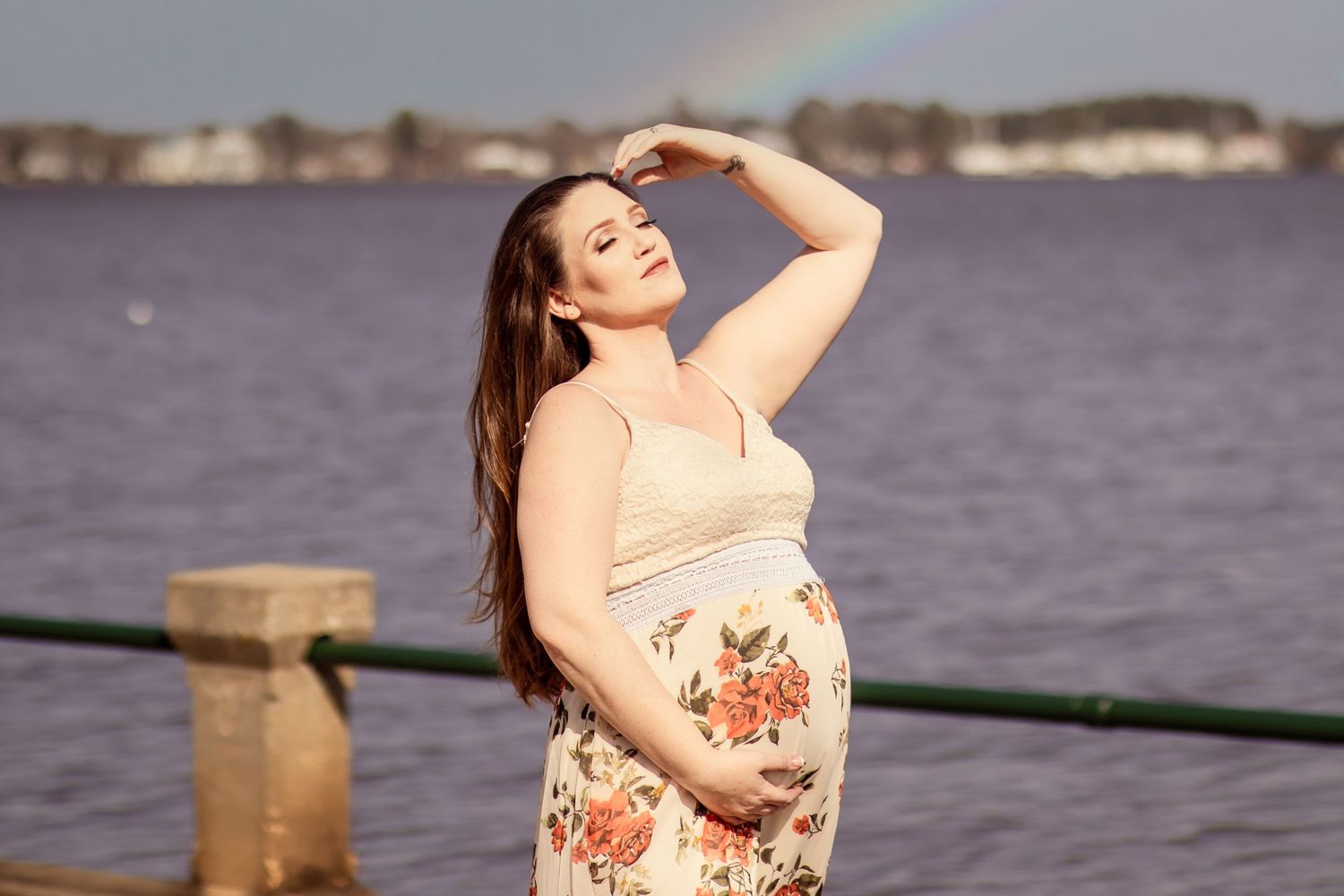 Glowing pregnant woman in floral outfit posing by lake with rainbow in sky.
