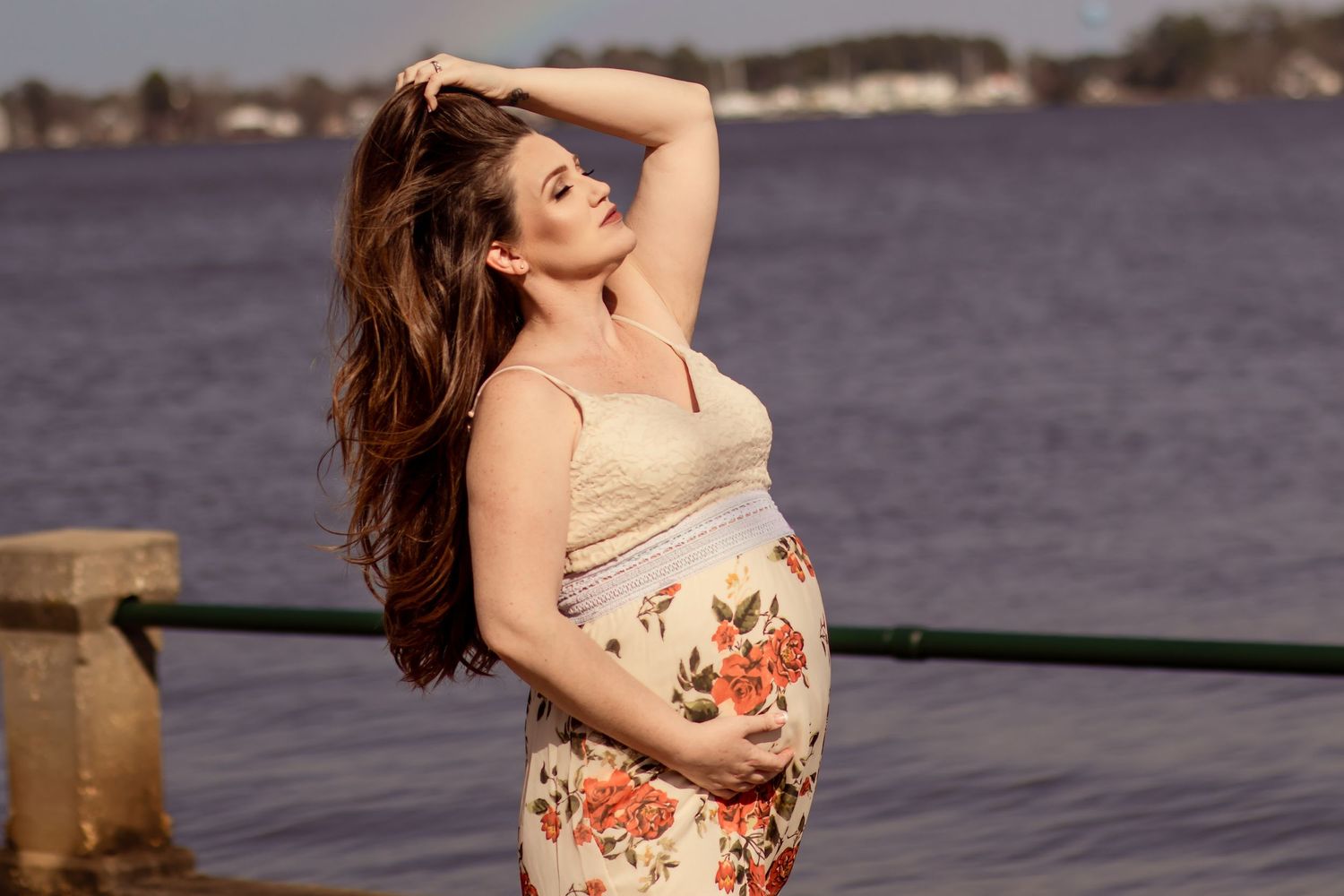 Pregnant woman in floral skirt holding belly near waterfront on sunny day.