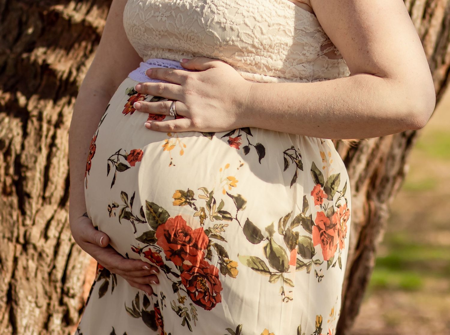 Close-up of pregnant woman in floral dress cradling baby bump beside rustic tree outdoors.