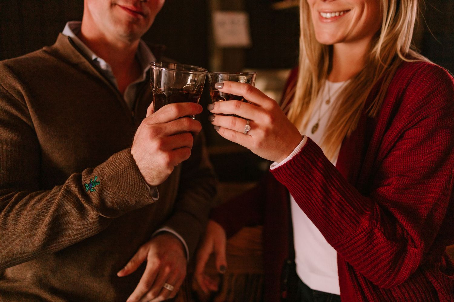 Couple clinking whiskey glasses in warm ambient light, celebrating together.
