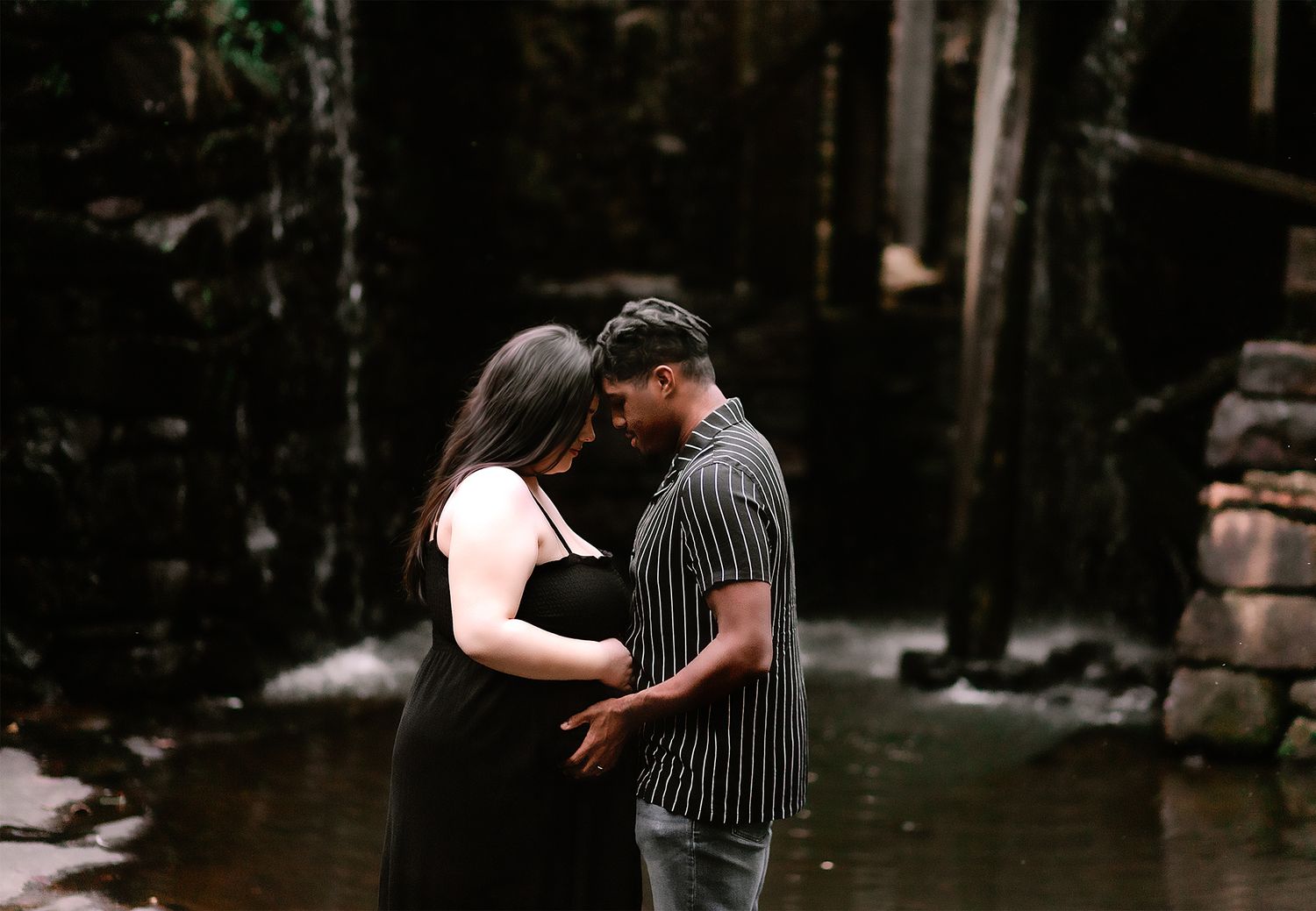 Expecting couple sharing an intimate forehead touch near a waterfall in a dramatic dark outdoor setting.