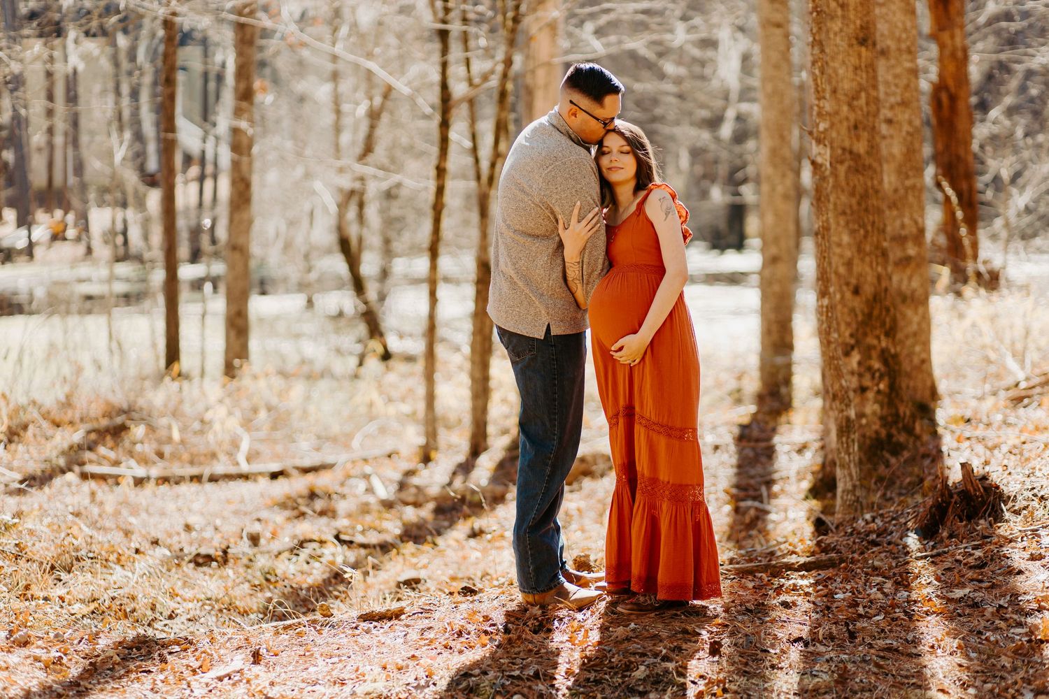 Expectant couple embracing outdoors in winter forest, woman in elegant rust red maternity gown with partner in grey sweater.