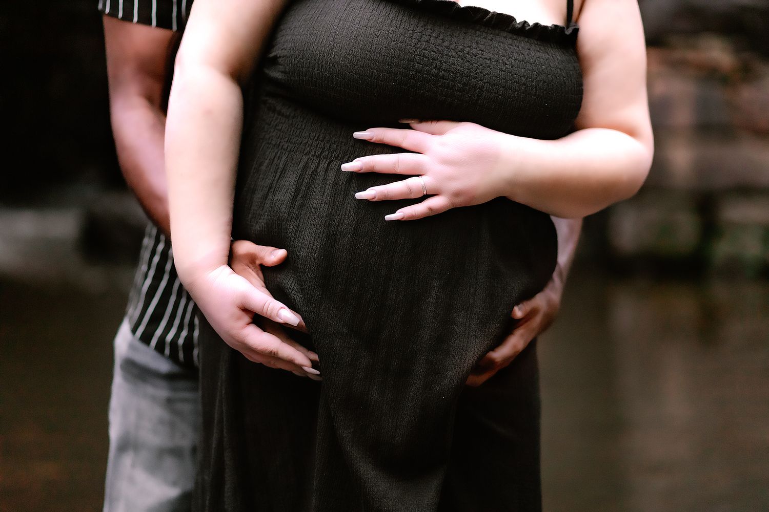 Pregnant woman in black dress cradling baby bump with partner embracing her from behind.