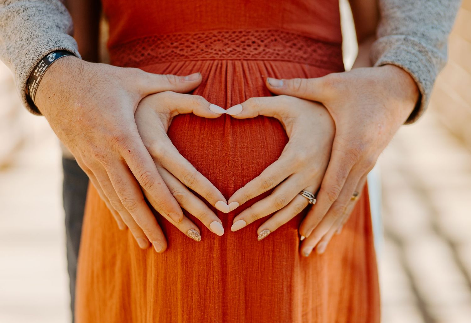 Couple forming a heart shape with their hands over a pregnant belly in a warm orange maternity photo.