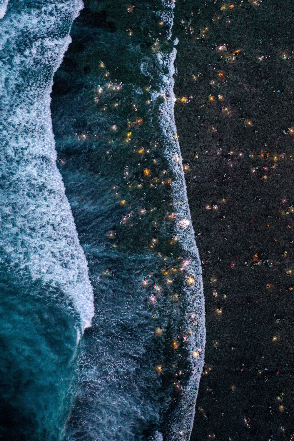 Aerial night view of coastline showing contrast between dark beach and illuminated city lights.