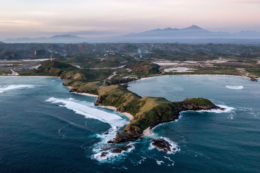Aerial view of a rugged coastal peninsula with waves crashing against cliffs and two curved bays during sunset.