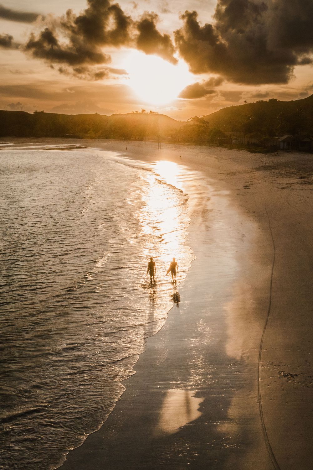 Two silhouettes walking on beach at sunset with golden light reflecting on wet sand.