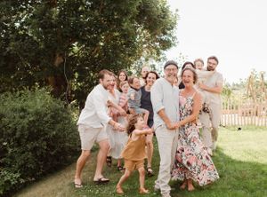 Extended family group poses playfully together in a sunny garden setting wearing coordinated light outfits.