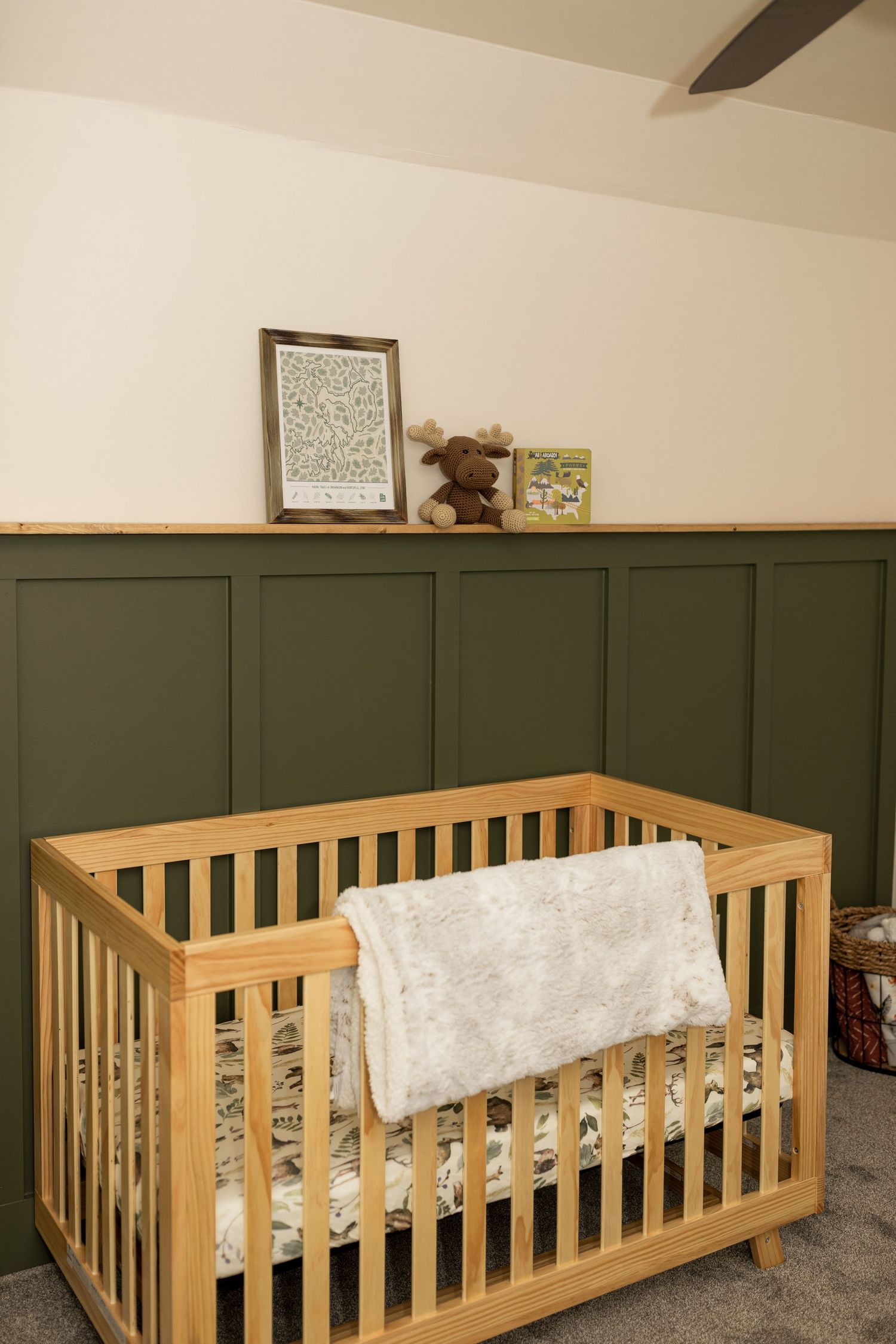Natural wood crib with white bedding against a dark green paneled accent wall in a nursery.