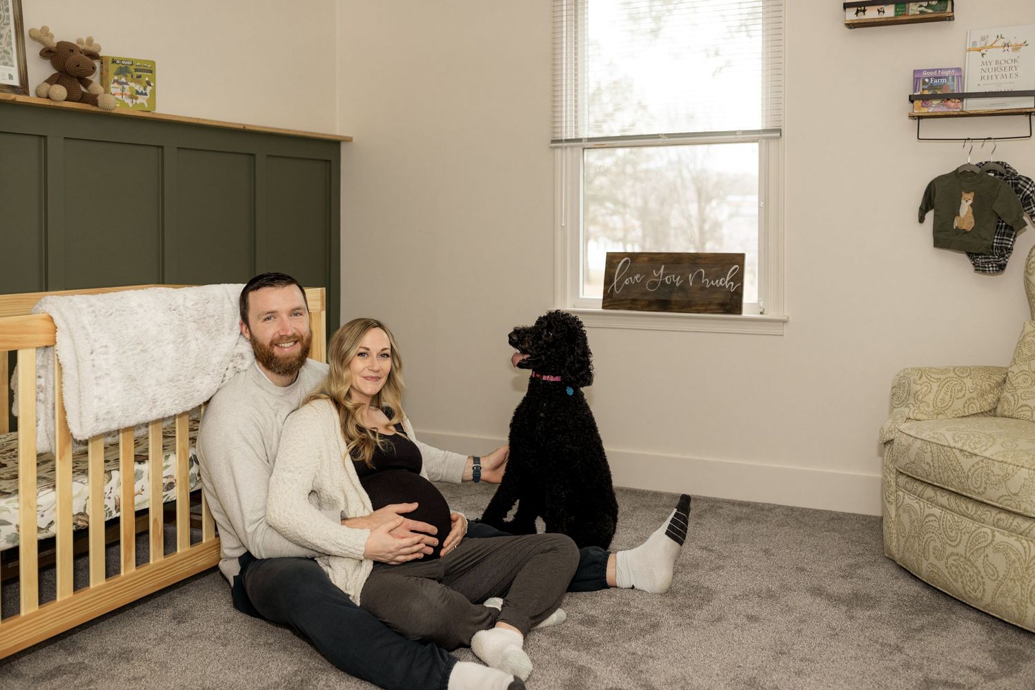 Wide view of nursery room showing crib, armchair, and people sitting with dog on floor.