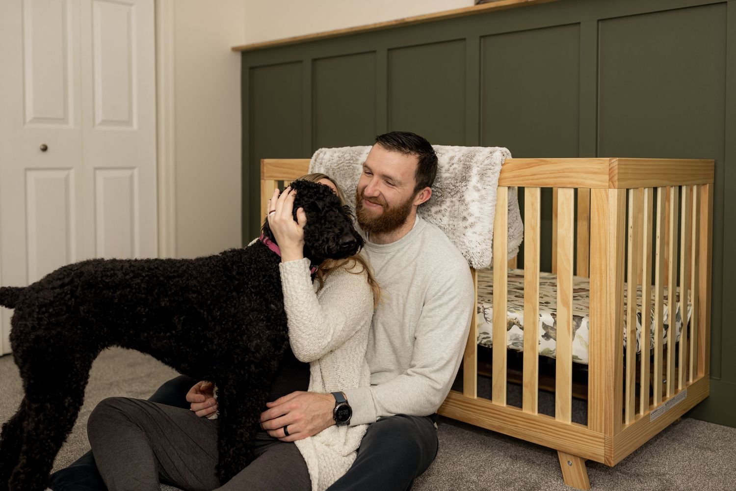 Family relaxes on carpet with their black dog near wooden furniture.