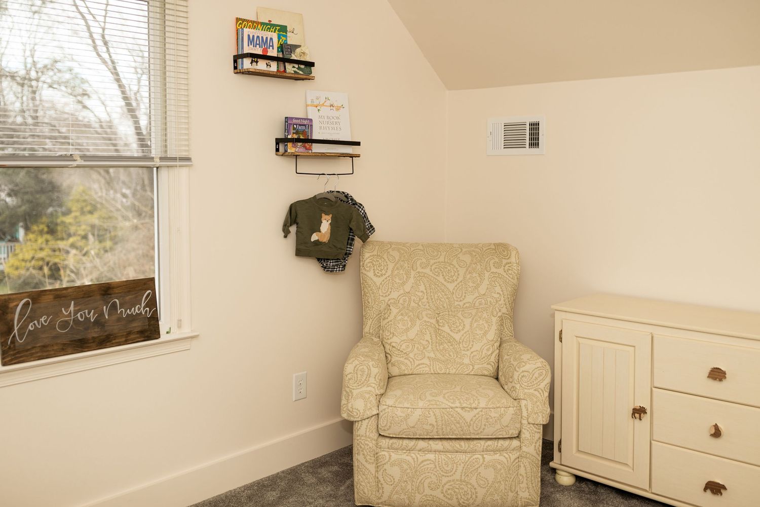 A modern nursery space featuring a beige armchair, wooden shelving, and wall decor.