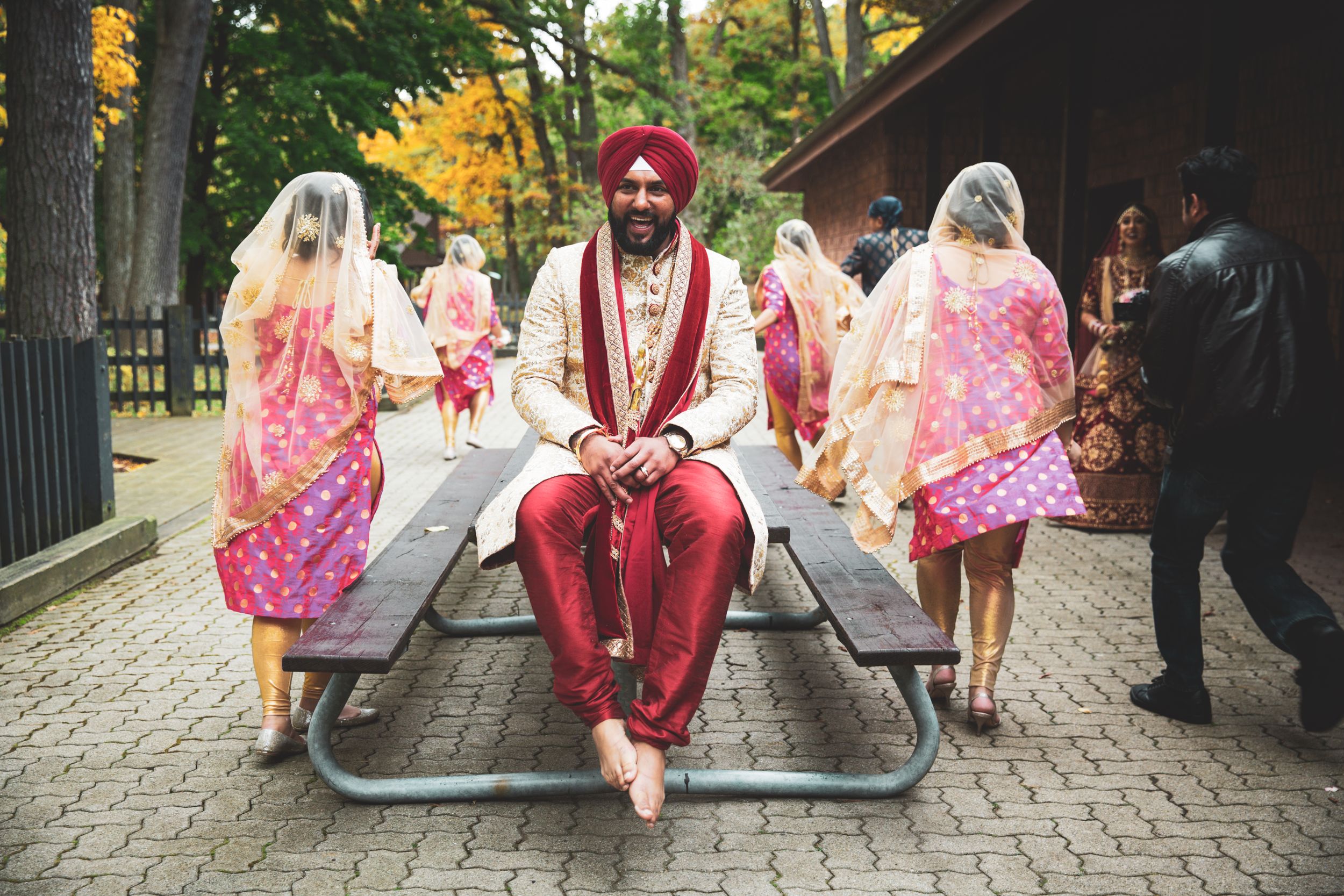 Wedding party scene with central figure in red turban surrounded by guests in pink attire outdoors.